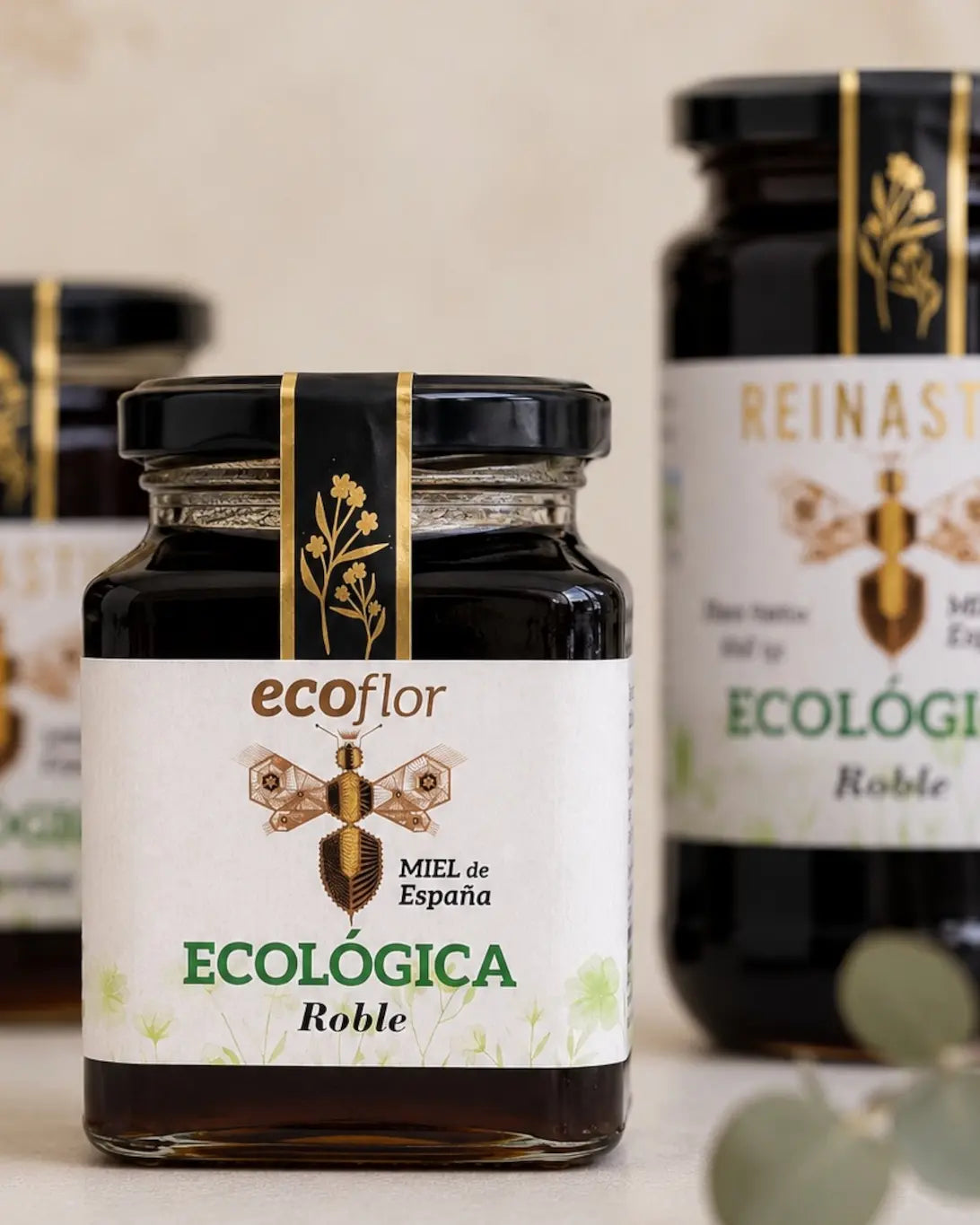 Primer plano de un tarro cuadrado de miel de Roble Ecoflor con dos botes grandes de Reinastur desenfocados al fondo.